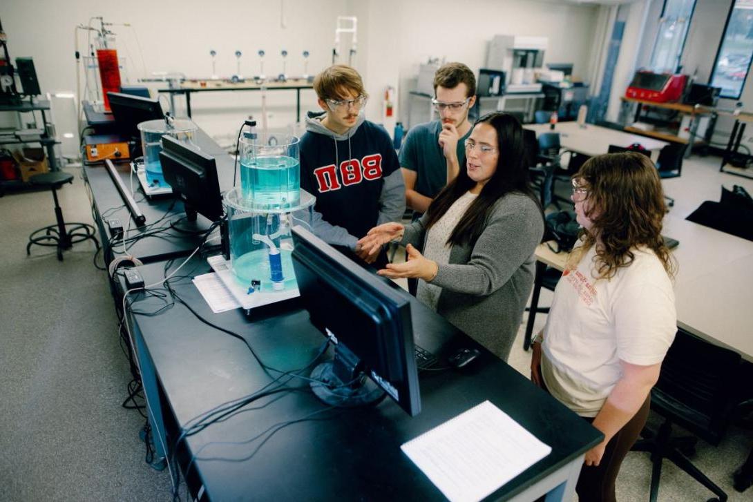 A Kettering professor instructs three students in the Chemical Engineering Lab. On the table is a computer and lab equipment.